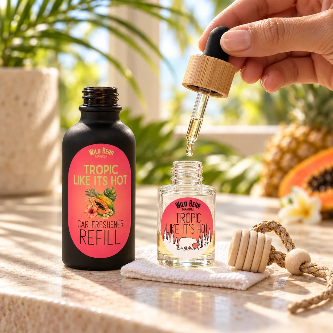 A hand uses a dropper to refill a Tropic Like It’s Hot glass car freshener beside its matching refill bottle and detached wooden lid.