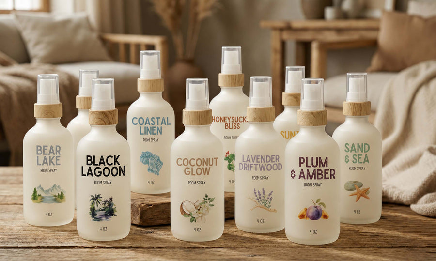 A group of ten room spray bottles from the brand 'Scent of the Sea' arranged on a wooden table in a cozy, softly lit living room setting. The frosted bottles feature light wood caps and are labeled with various scents including Black Lagoon, Coconut Glow, Lavender Driftwood, Plum & Amber, and Sand & Sea.